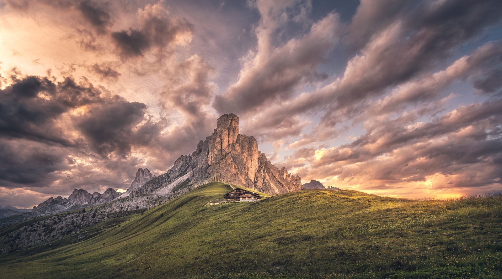 Pradera en primer plano, un refugio y una montaña al fondo bajo nubes iluminadas.